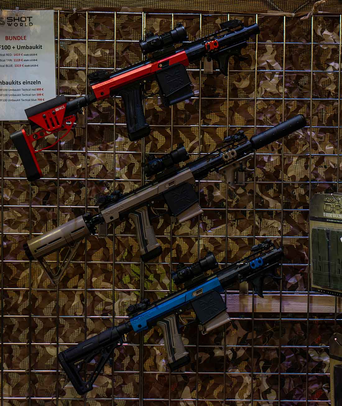 An einer Wand mit Tarnnetzen hängen drei bunte Paintballmarkierer